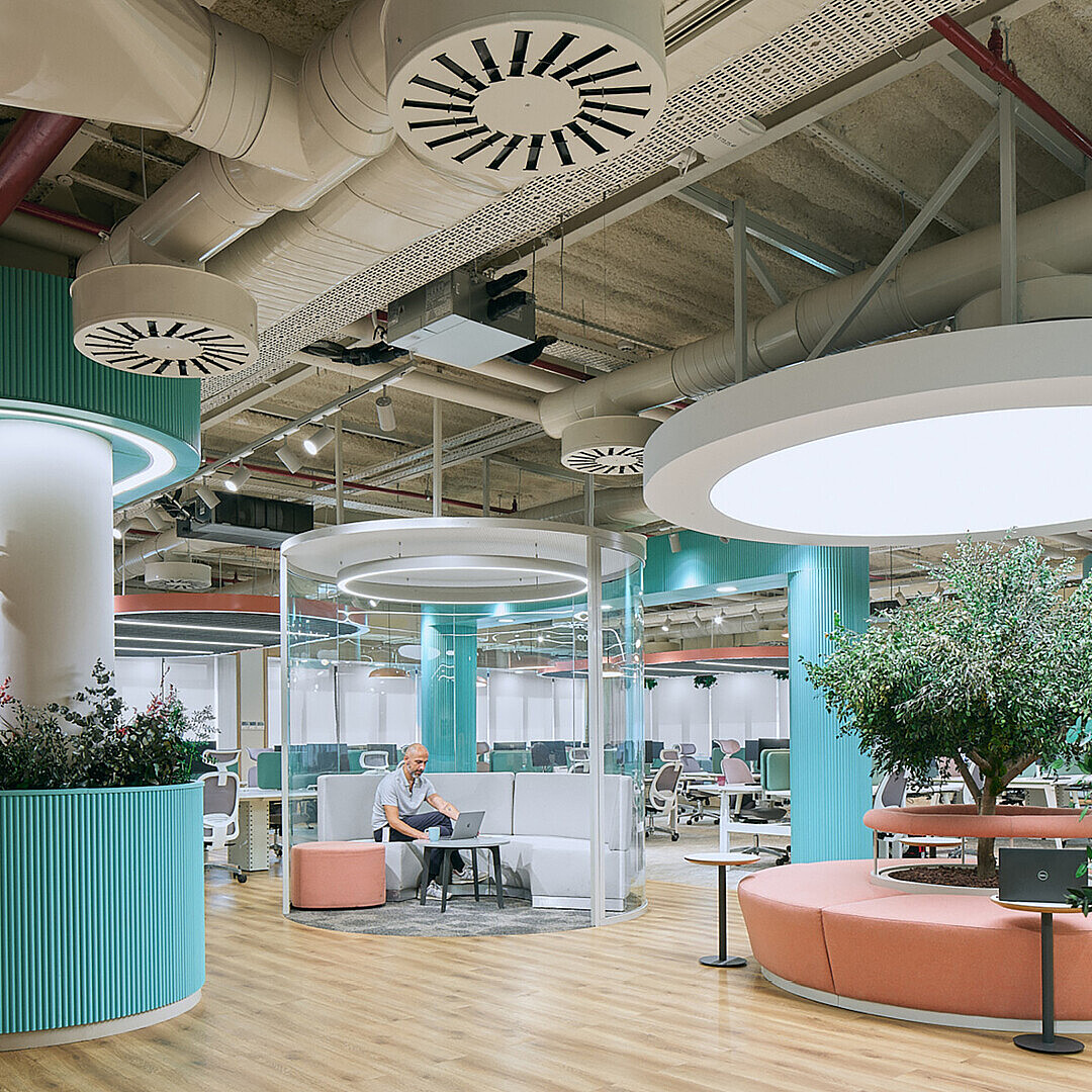 Open-plan office space with circular seating areas, large round ceiling lights, plants, and a man working on a laptop inside a transparent cylindrical booth