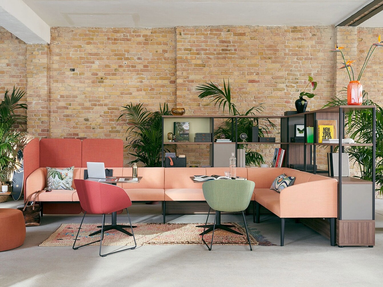 Salon ouvert avec un canapé modulable couleur pêche, deux chaises modernes rouges et vertes, des tables basses, des plantes et une étagère contre un mur en briques