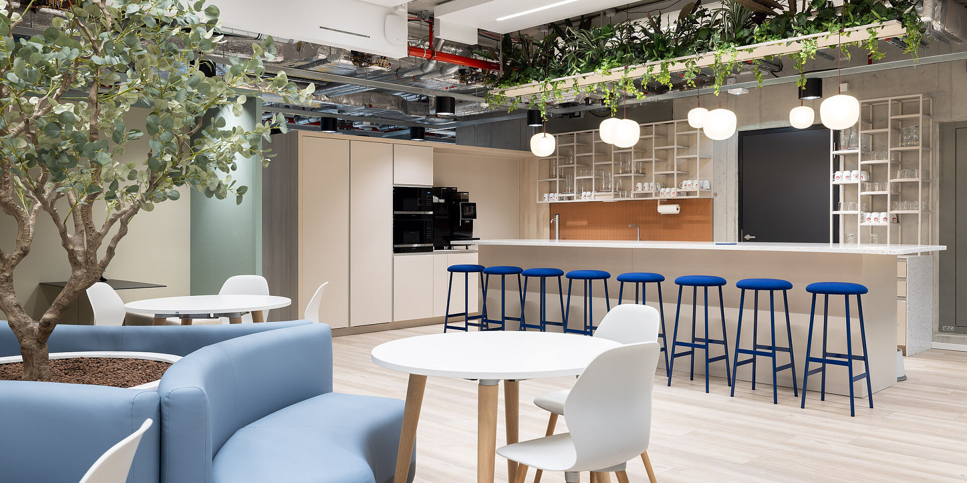 Moderna zona de bistró en Veolia con sillas de diseño se:spot en blanco y taburetes de bar azules en mesas se:assist, rodeadas de plantas y una iluminación clara. 