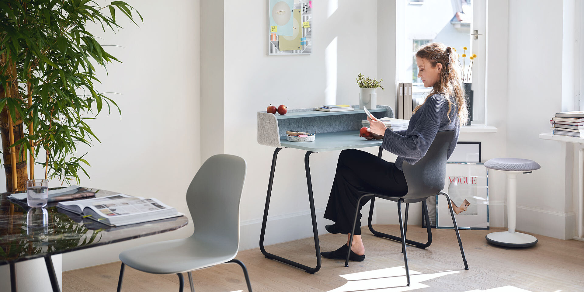 Frau sitzt an einem modernen Schreibtisch mit schwarzen Beinen und grauer Ablage, liest Dokument in hellem, minimalistischem Arbeitszimmer mit Pflanzen und großen Fenstern