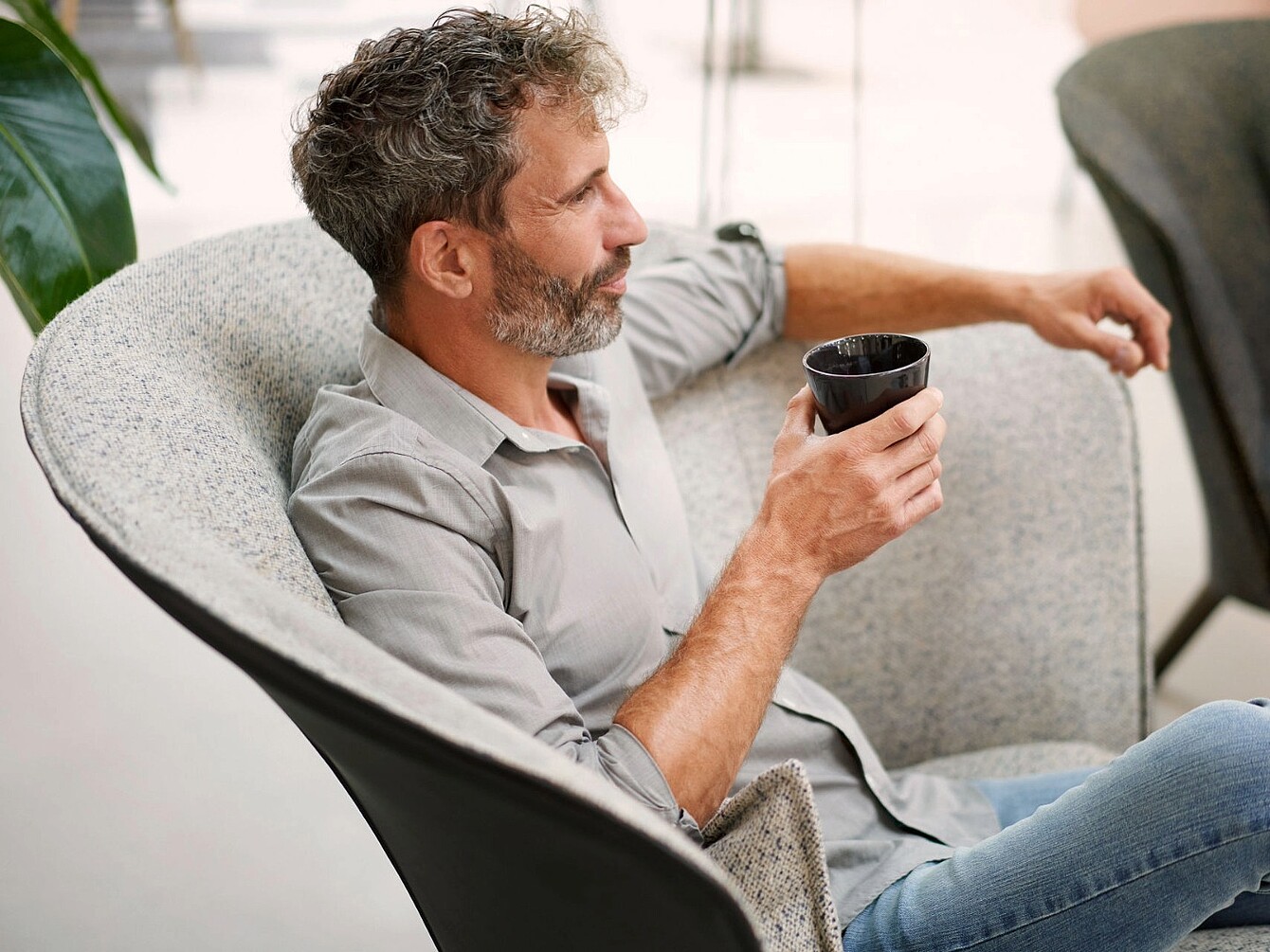 Hombre con barba sentado en un sillón curvo, sosteniendo una taza negra y mirando hacia un lado