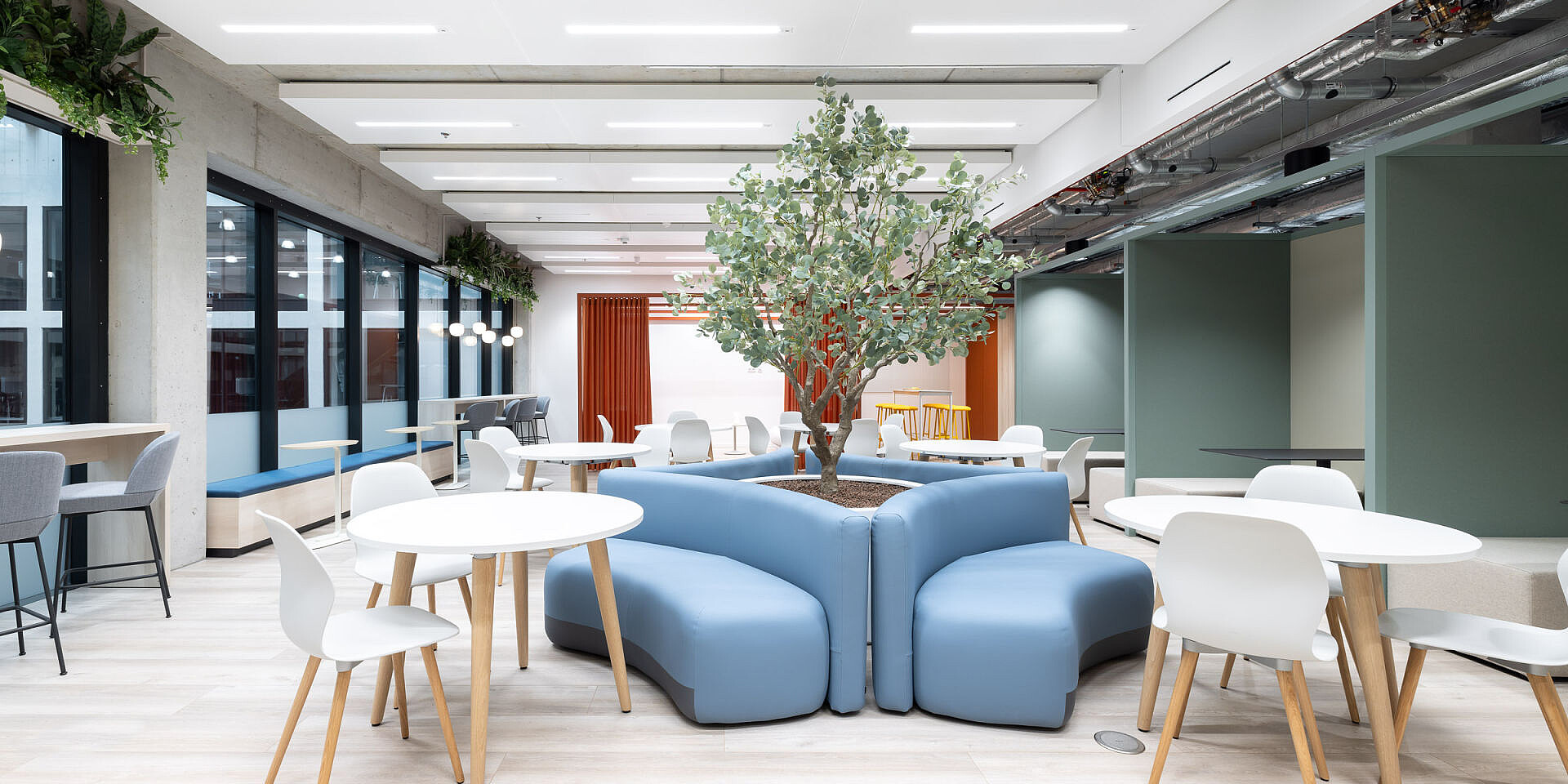 Cantina luminosa con mesas redondas blancas, sillas de diseño se:spot azules y un árbol central en un asiento azul, mobiliario moderno en Veolia.  