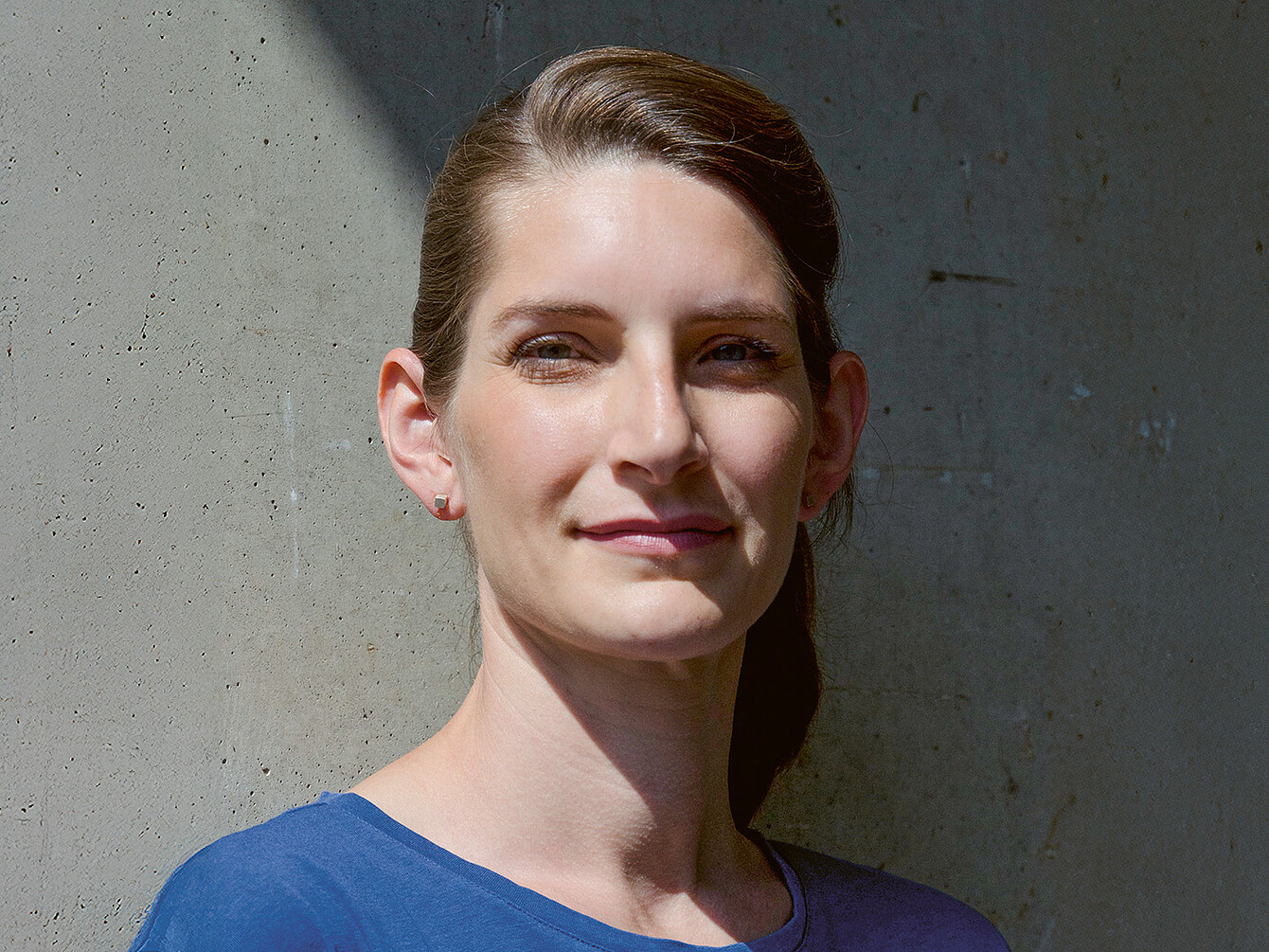 Portret van industrieel ontwerper Judith Daur, zelfverzekerd glimlachend tegen een betonnen achtergrond met textuur, met een blauw topje aan.