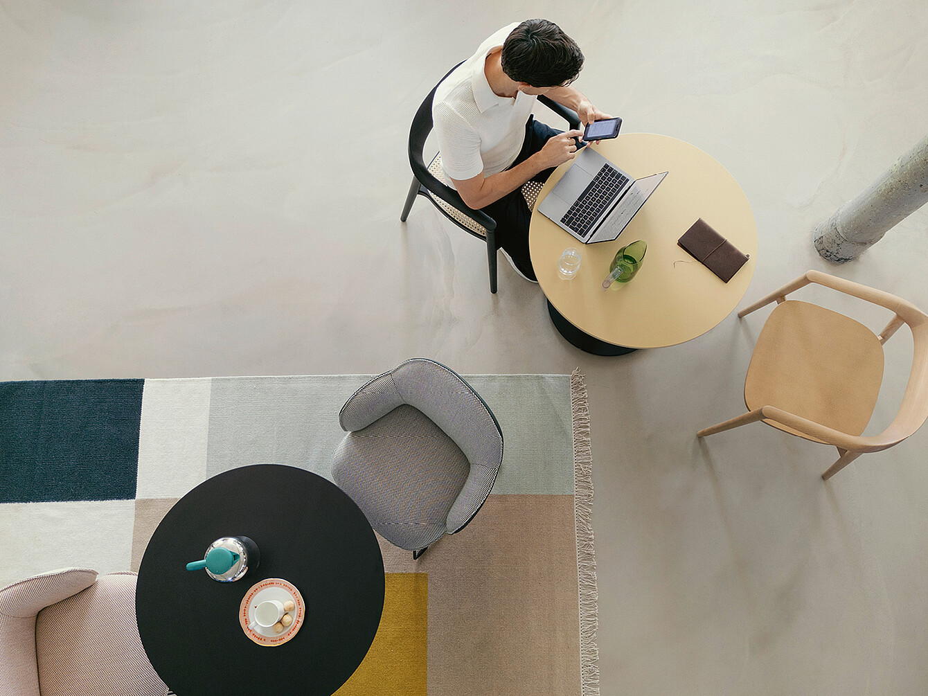 personne assise à une table ronde dans un Work Café Bibliothèque de bureau moderne, utilisant un smartphone et un ordinateur portable, entourée de chaises et d’un tapis, illustrant un refuge calme pour un travail concentré.