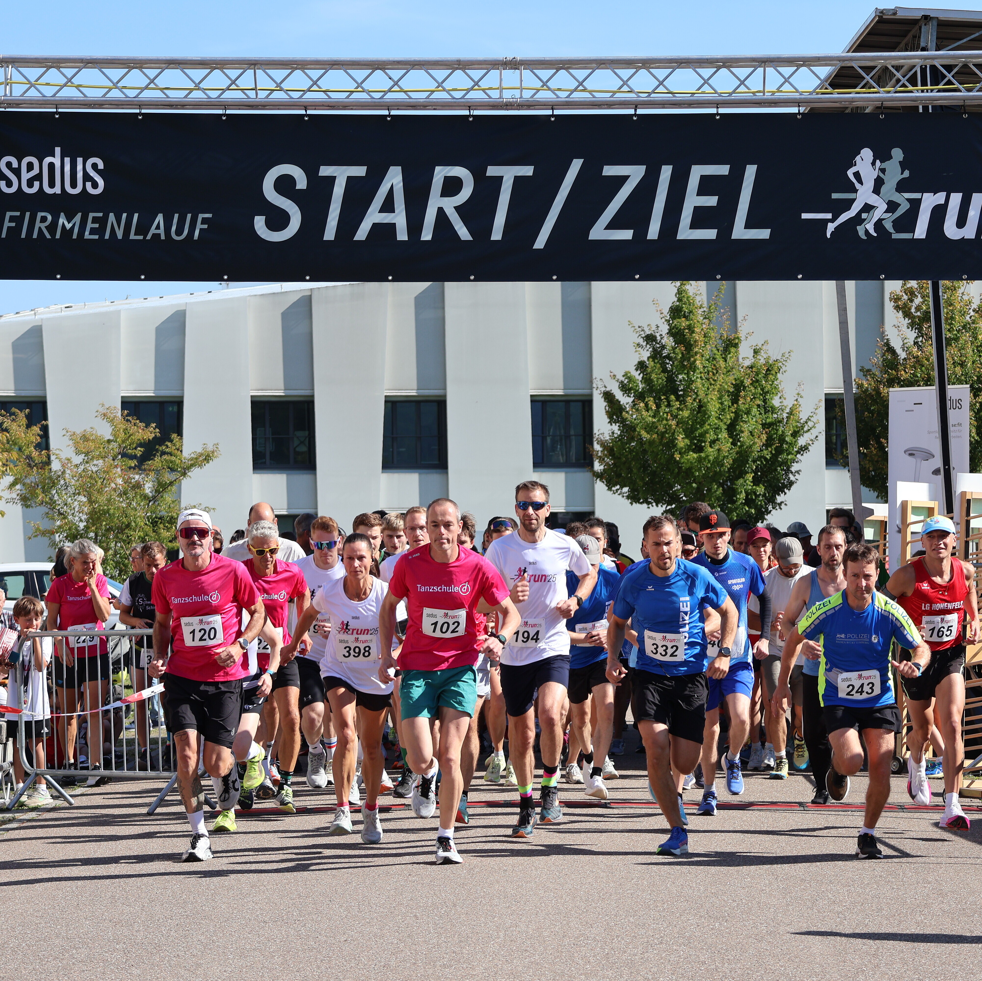 Läufer starten beim Sedus Firmenlauf 2025 unter dem Banner 'START / ZIEL - run' bei sonnigem Wetter auf asphaltierter Strecke vor modernem Gebäude.