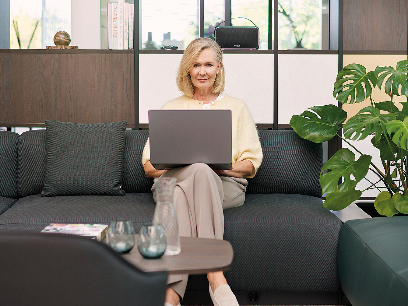 Une femme aux cheveux blonds mi-longs est assise sur un canapé moderne se:living gris foncé de Sedus, un ordinateur portable ouvert posé sur ses genoux. Elle porte un chemisier clair et un pantalon beige. À l’arrière-plan, on aperçoit le système d’étagères se:matrix de Sedus orné d’objets décoratifs, une grande plante d’intérieur aux feuilles caractéristiques, ainsi qu’une fenêtre donnant sur des arbres. Sur la table basse devant elle se trouvent une carafe d’eau, deux verres et un magazine.