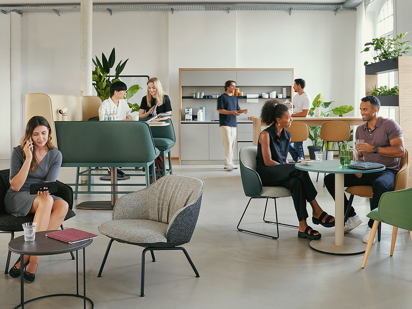 Een modern Work Café met verschillende zitopstellingen. Mensen zijn in gesprek, sommigen gebruiken hun telefoon. Er zijn groene planten te zien en op de achtergrond is een kitchenette te zien.