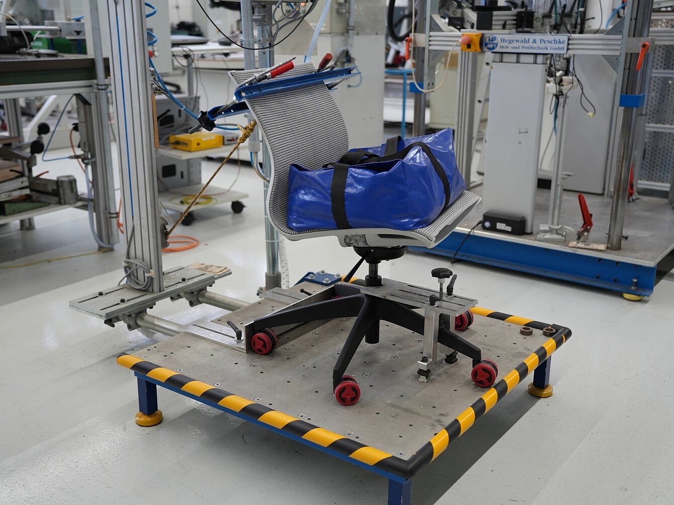 Bureaustoel met rugleuning van gaas en blauwe tas op een testplatform met geel-zwart gestreepte randen in een laboratoriumomgeving