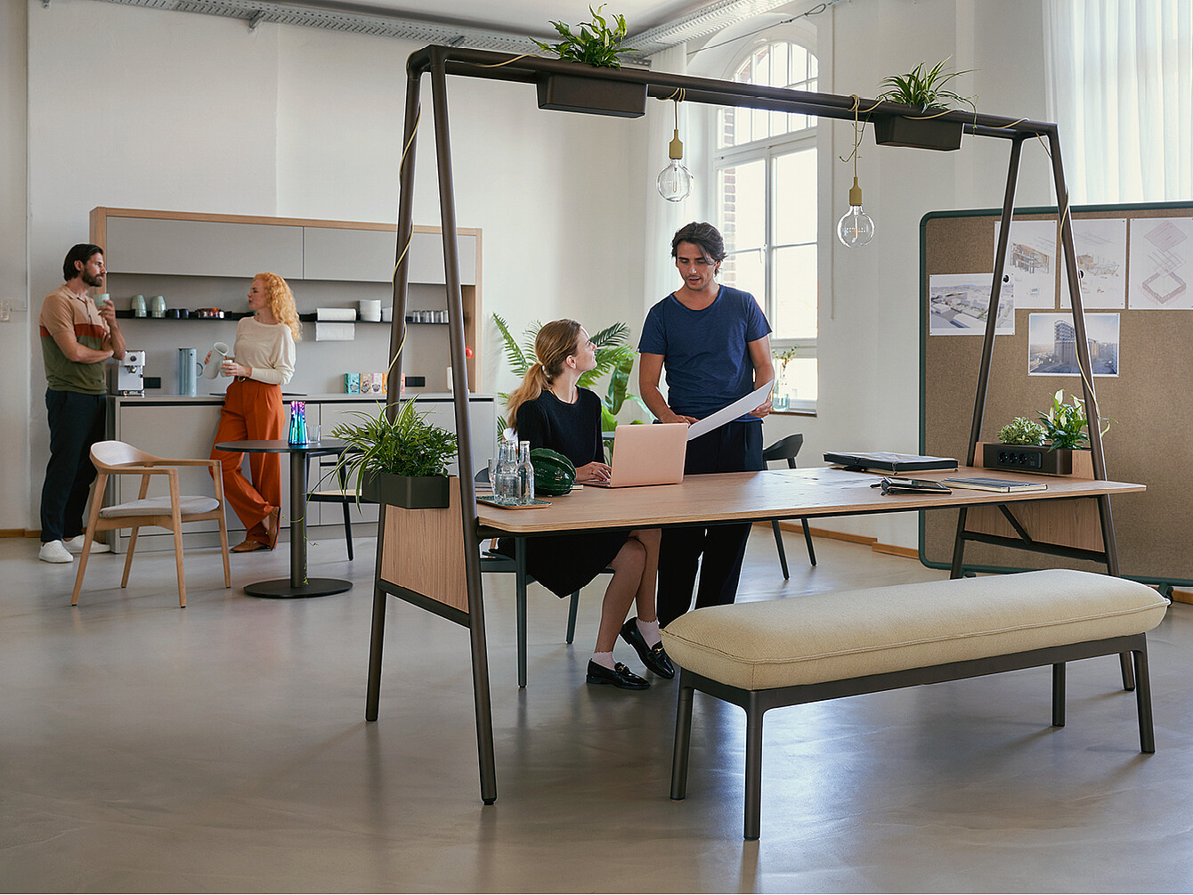 Moderne hybride duktile Gemeinfläche mit zwei Personen am Tisch, die an Laptop und Papier arbeiten, und zwei Personen im Hintergrund an der Kaffeeküche im offenen, hellen Büro mit Pflanzen und Pinnwand