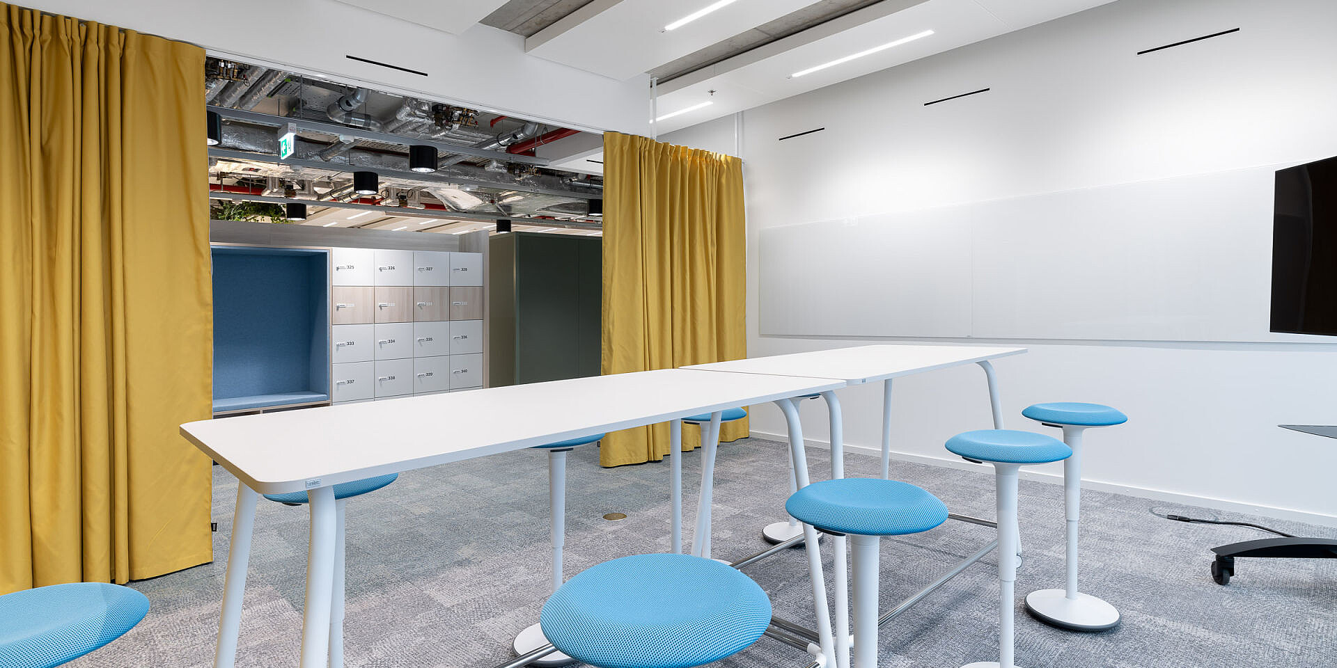 Moderna sala de reuniones con mesa de taller se:lab high desk blanca, rodeada de taburetes se:fit azules y taburetes se:dot, cortinas amarillas y paredes blancas. 