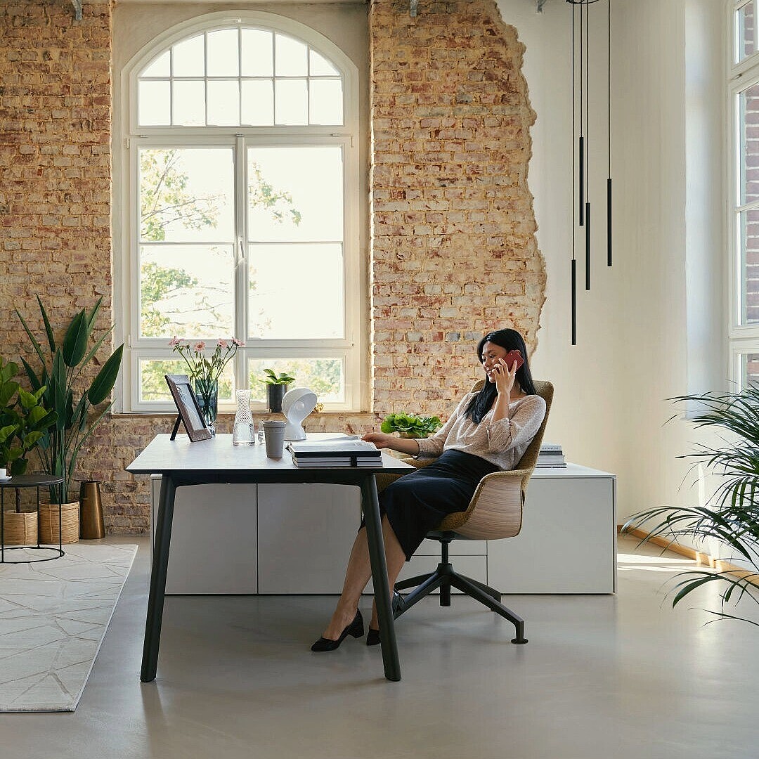 Moderne Büroeinrichtung mit großem Fenster, Pflanzen, Sitzgruppe und einer Person am Schreibtisch mit Laptop und Telefon