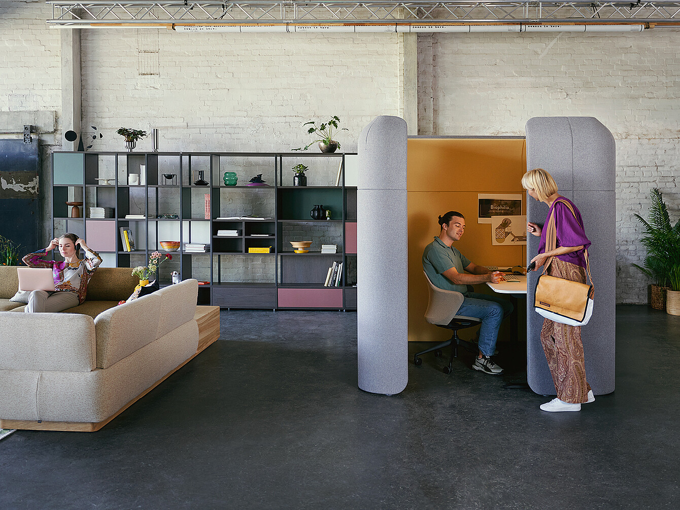 Een modern Work Café met een gezellige zithoek met een grijze bank en een persoon die op een laptop werkt. Op de voorgrond staat een vrouw in een paarse top en een rok met patroon naast een geluiddichte cabine waar een persoon aan een bureau zit.