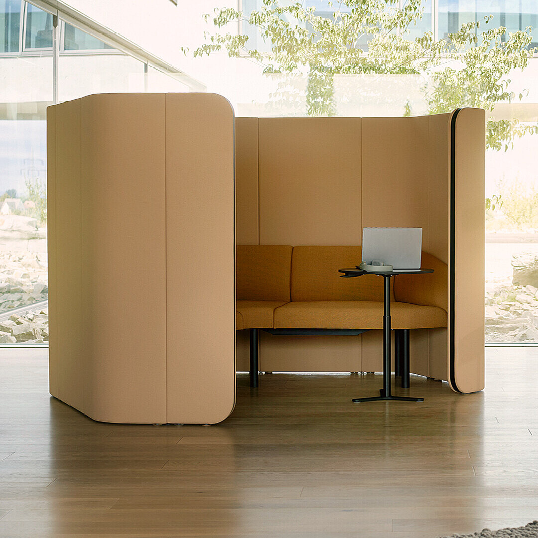 Abgeschirmte Sitznische mit Sofa, kleinem Tisch und Laptop in einer hellen, modernen Büroumgebung.