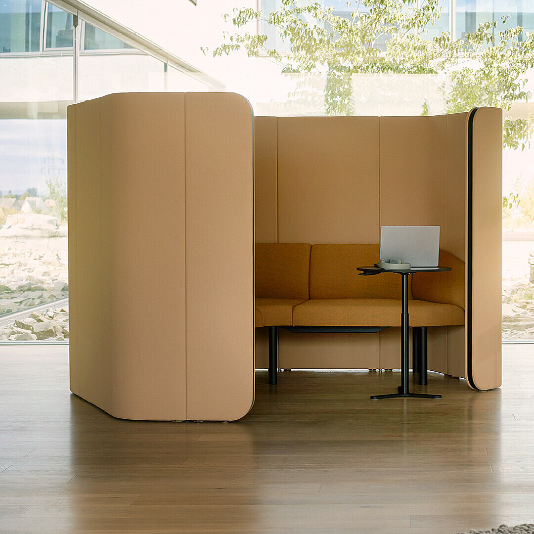 Modern office space with a high-backed brown booth containing a laptop on a small black table, a coat rack with hanging items, and light-colored armchairs around a low table with two cups