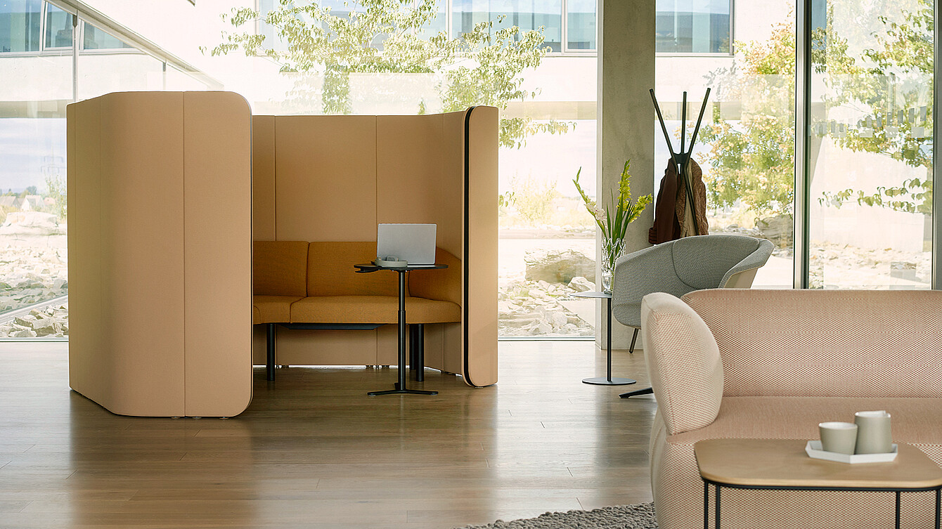 Cabina de asiento aislada con respaldo alto y paneles laterales, sofá, mesa pequeña y ordenador portátil en un entorno de oficina luminoso.