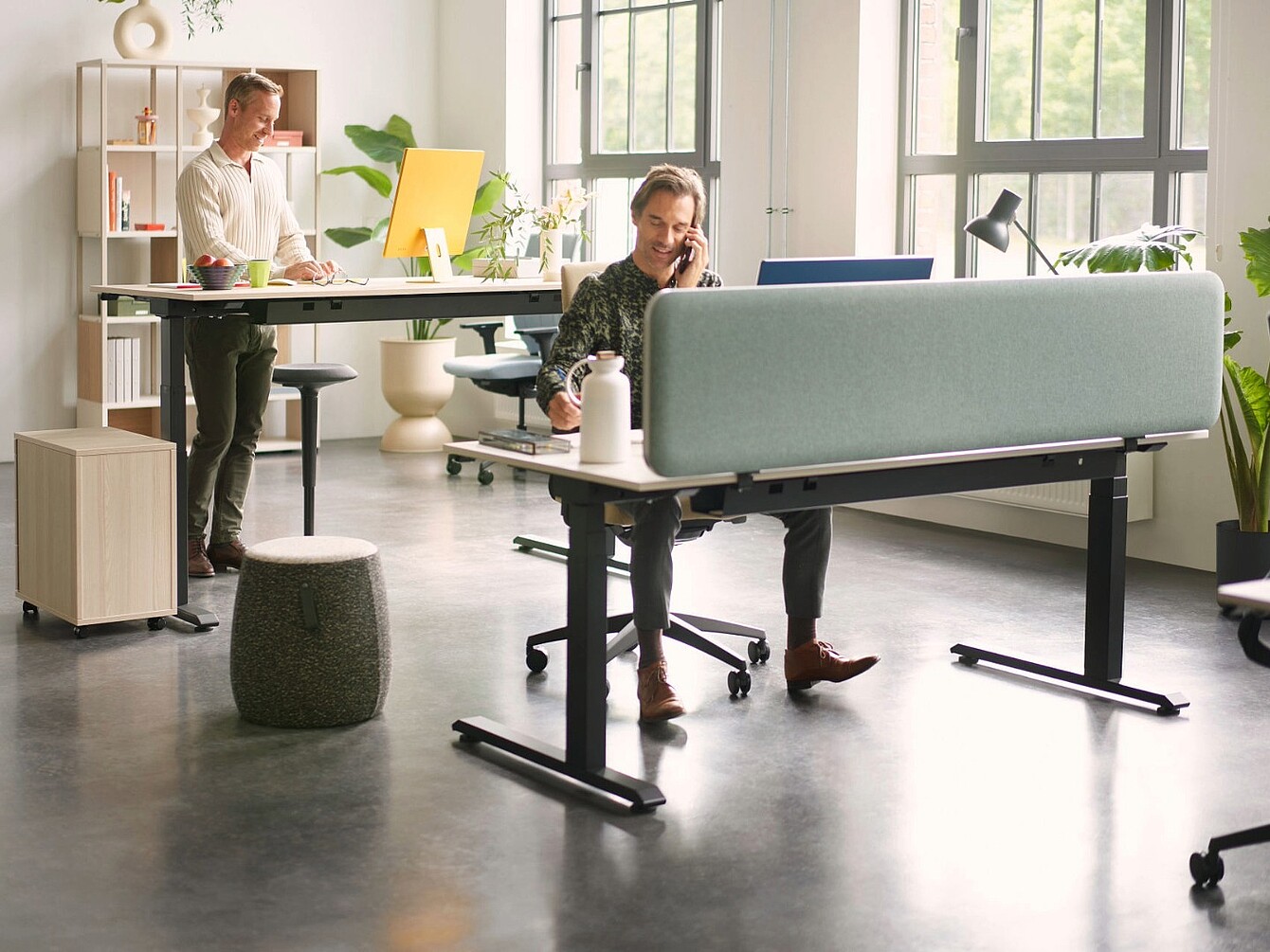 Spazio ufficio moderno con due persone che lavorano a scrivanie separate da un pannello divisorio per la privacy azzurro chiaro, con sedie ergonomiche, una scrivania per lavorare in piedi e luce naturale proveniente da grandi finestre