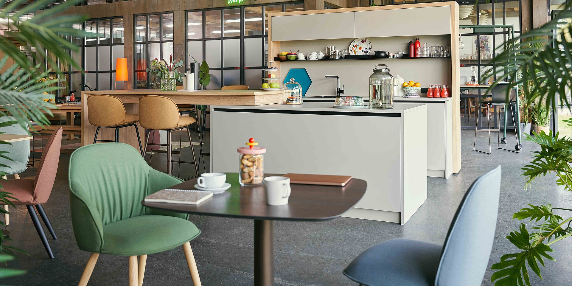 Een moderne gemeenschappelijke ruimte met een kleine keuken, een tafel met groene en roze stoelen op de voorgrond en extra zitgelegenheden op de achtergrond. Planten en warme verlichting zorgen voor een uitnodigende sfeer.