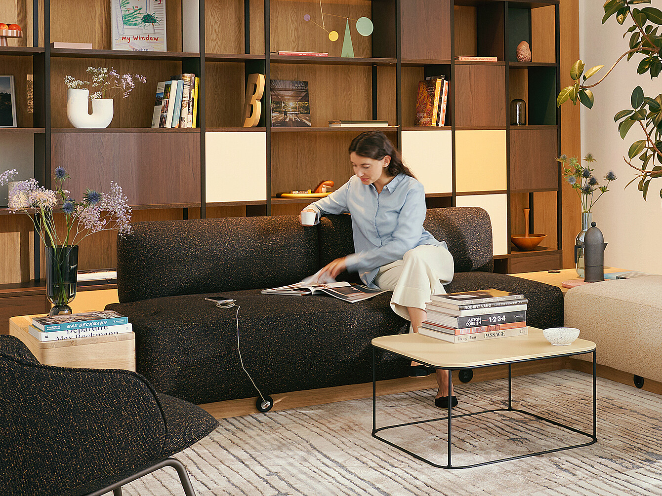 Personne assise sur un canapé sombre dans un Work Café , lisant un magazine avec une tasse de café, entourée d’étagères et de meubles modernes