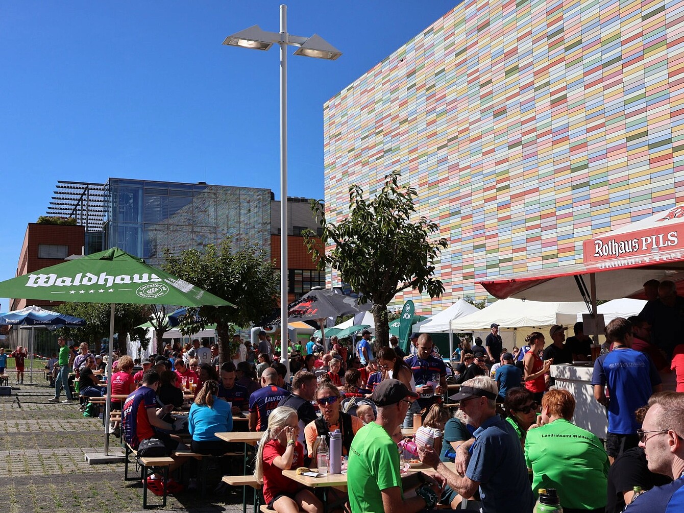 Menschen sitzen bei Sonnenschein an Biergartenbänken unter Schirmen mit den Aufschriften 'Waldhaus' und 'Rothaus Pils' vor einem bunten, gestreiften Gebäude.