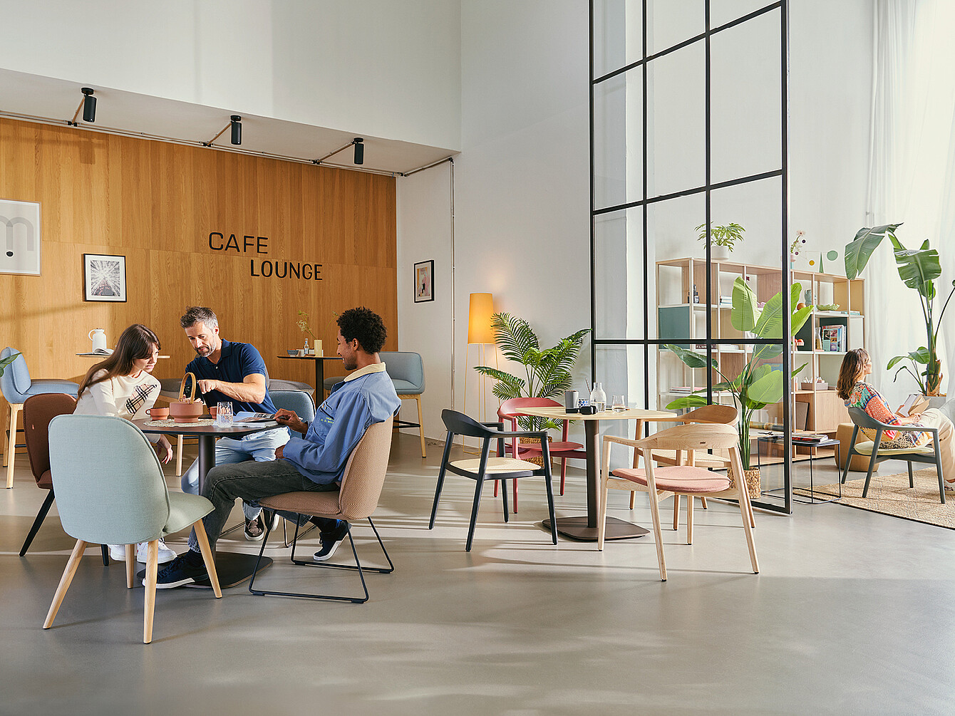 Trois personnes assises à une table dans un café lounge moderne avec des murs en bois, des plantes et divers sièges, dont des fauteuils et des tabourets