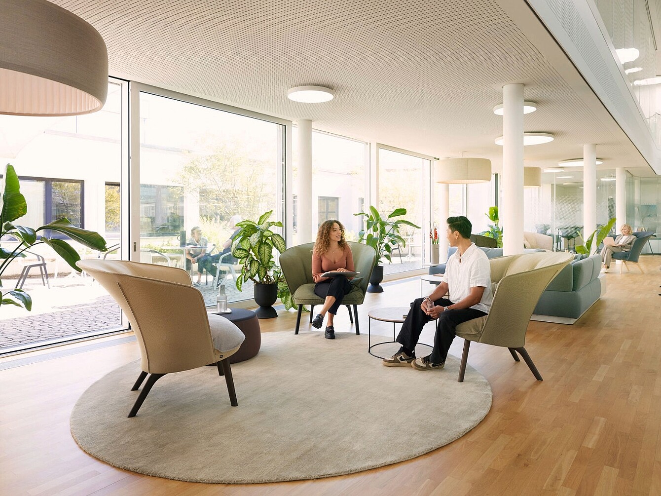 Dos personas sentadas en sillones beige alrededor de una pequeña mesa redonda sobre una alfombra circular, en una sala espaciosa y bien iluminada con grandes ventanas y plantas