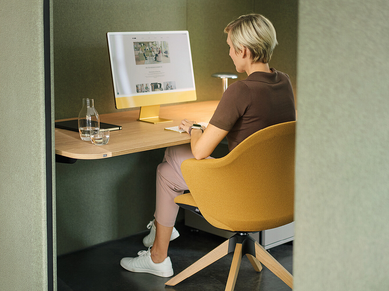 Une personne aux cheveux blonds courts est assise à un bureau moderne dans un espace de travail séparé et insonorisé. Elle porte un t-shirt marron, un pantalon beige et des baskets blanches. La chaise de bureau est jaune et de forme arrondie. Sur le bureau se trouvent un grand ordinateur avec un socle jaune, un verre d’eau et un cahier. Les murs de l’espace de travail sont recouverts d’un tissu verdâtre, créant une atmosphère apaisante.
