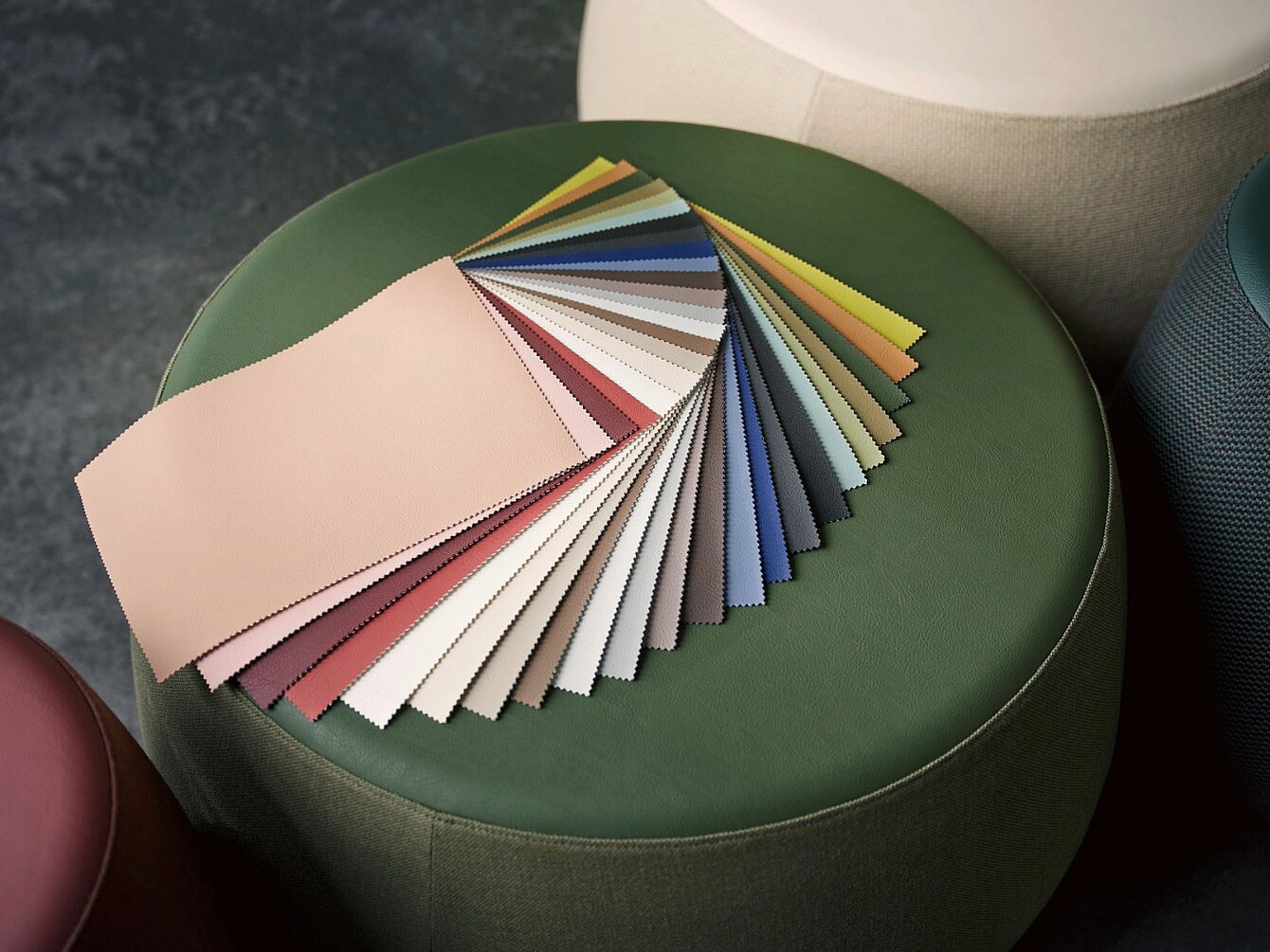 Fan of fabric swatches in various colors arranged on a round green ottoman with parts of other ottomans visible around it