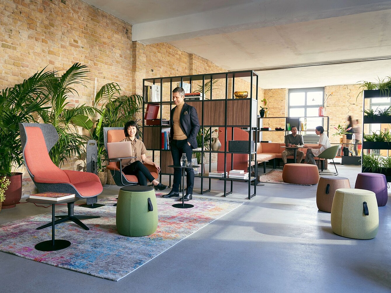 Salon ouvert avec des sièges modernes, notamment des fauteuils, des tabourets et un canapé modulaire, séparés par une étagère en métal noir et en bois, avec en toile de fond un mur en briques, de grandes fenêtres et des plantes d’intérieur.