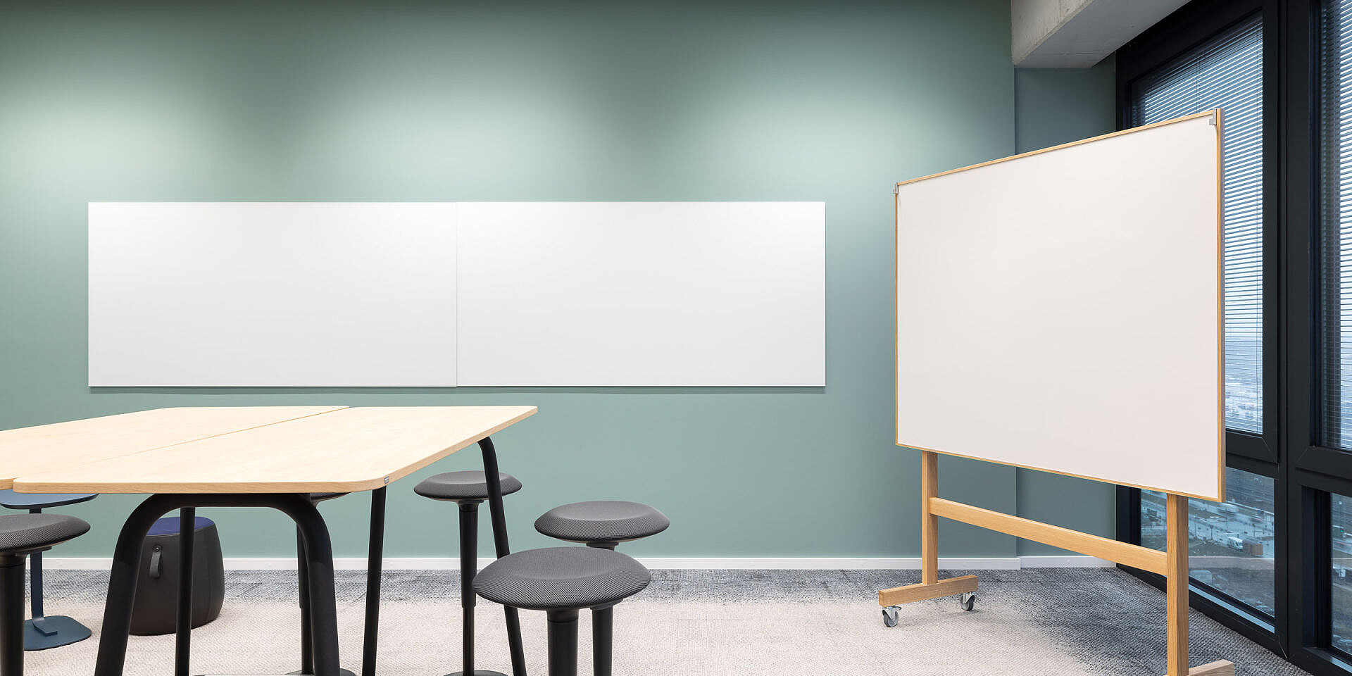 Área de trabajo moderna con mesa de taller se:lab high desk, asientos de pie se:fit y varios taburetes universales se:dot delante de una pared verde y una pizarra blanca móvil. 