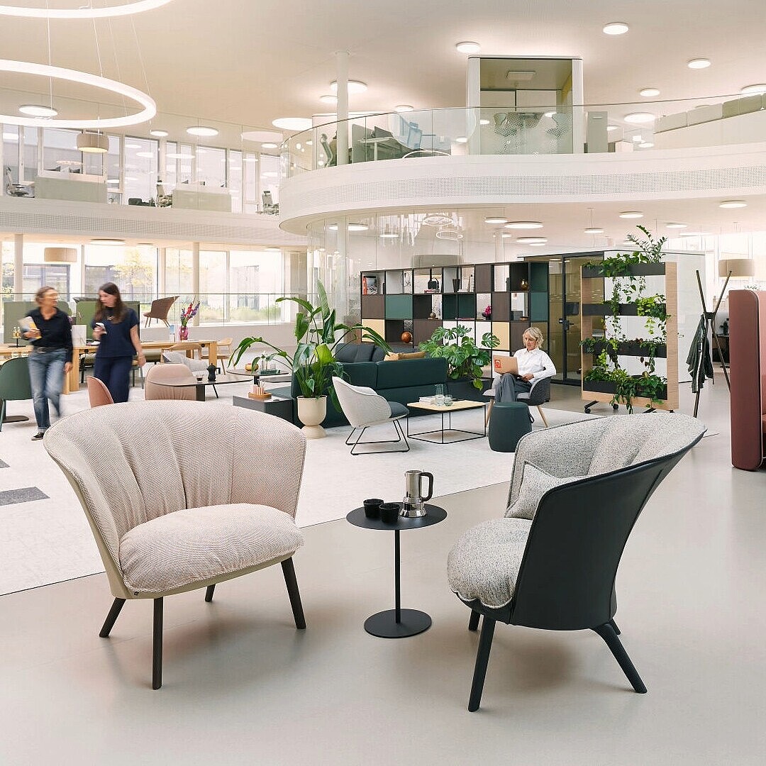 Open office space with two armchairs and a small table in the foreground and a booth with two people seated and talking in the background