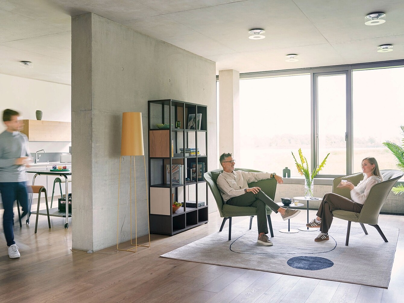 Dos personas sentadas en sillones modernos, una frente a la otra, en una sala luminosa con grandes ventanales, una alfombra, una lámpara de pie y una estantería, con una persona difuminada caminando al fondo cerca de la zona de la cocina