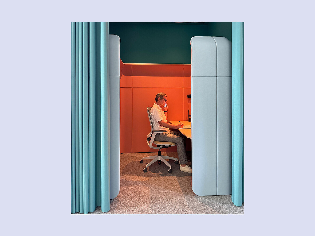 Persoon zittend op een bureaustoel, werkend aan een bureau in een kleine, afgesloten werkruimte met beklede wanden en een gordijn aan één kant