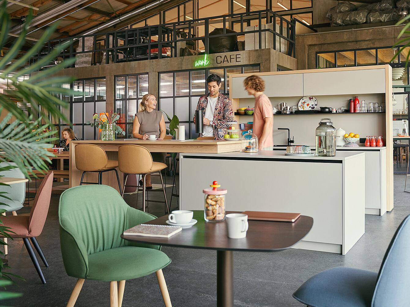 Bureau moderne Work Café avec des personnes discutant autour d’un îlot de cuisine, avec des tabourets de bar, des tasses à café, des biscuits et une pancarte « Work Café ».