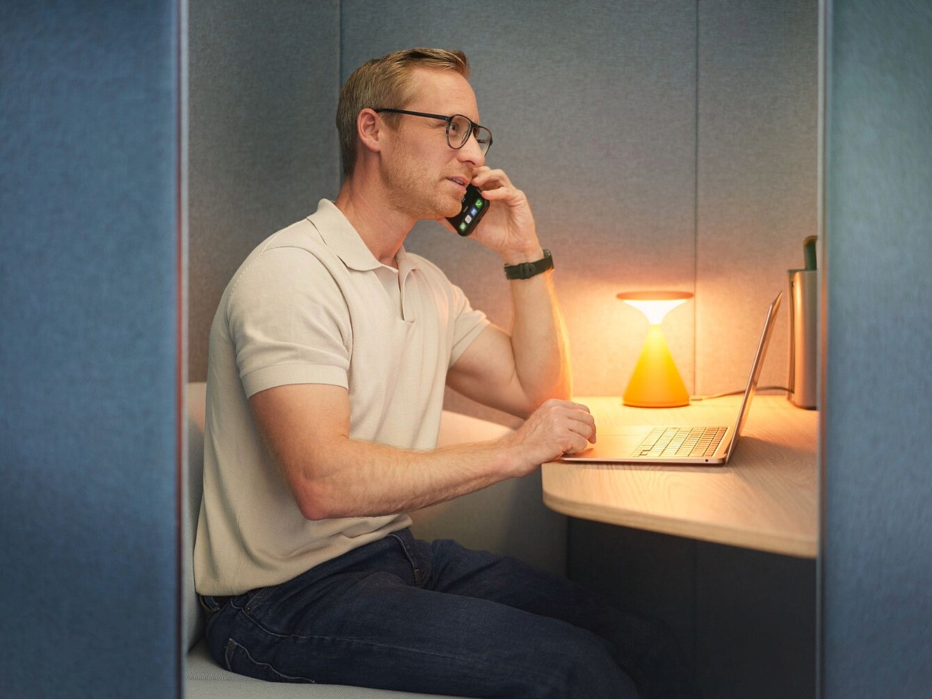 Hombre con gafas sentado en una pequeña cabina de oficina hablando por teléfono, con un ordenador portátil y una pequeña lámpara sobre la mesa