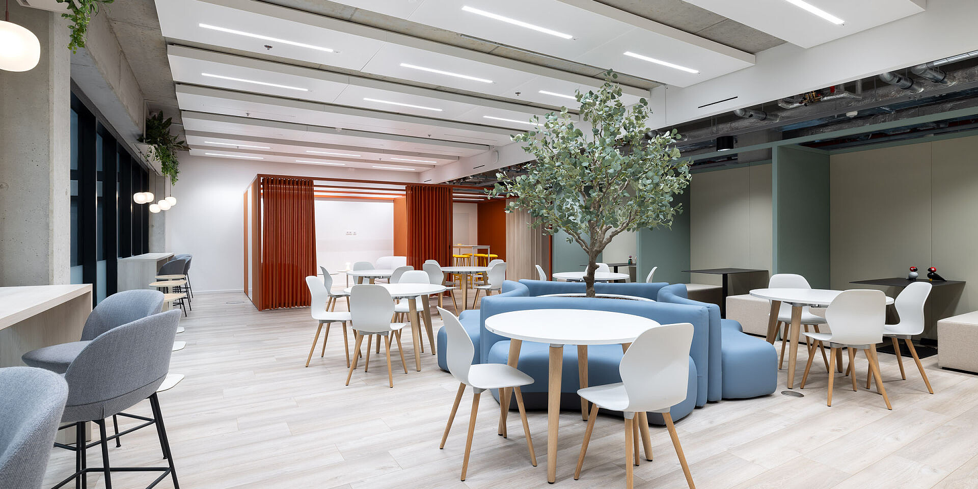 Moderna zona de bistró en Veolia con mesas redondas se:assist y sillas de diseño ergonómicas se:spot en colores claros, rodeadas de sofás y una gran planta para descansar relajadamente.  