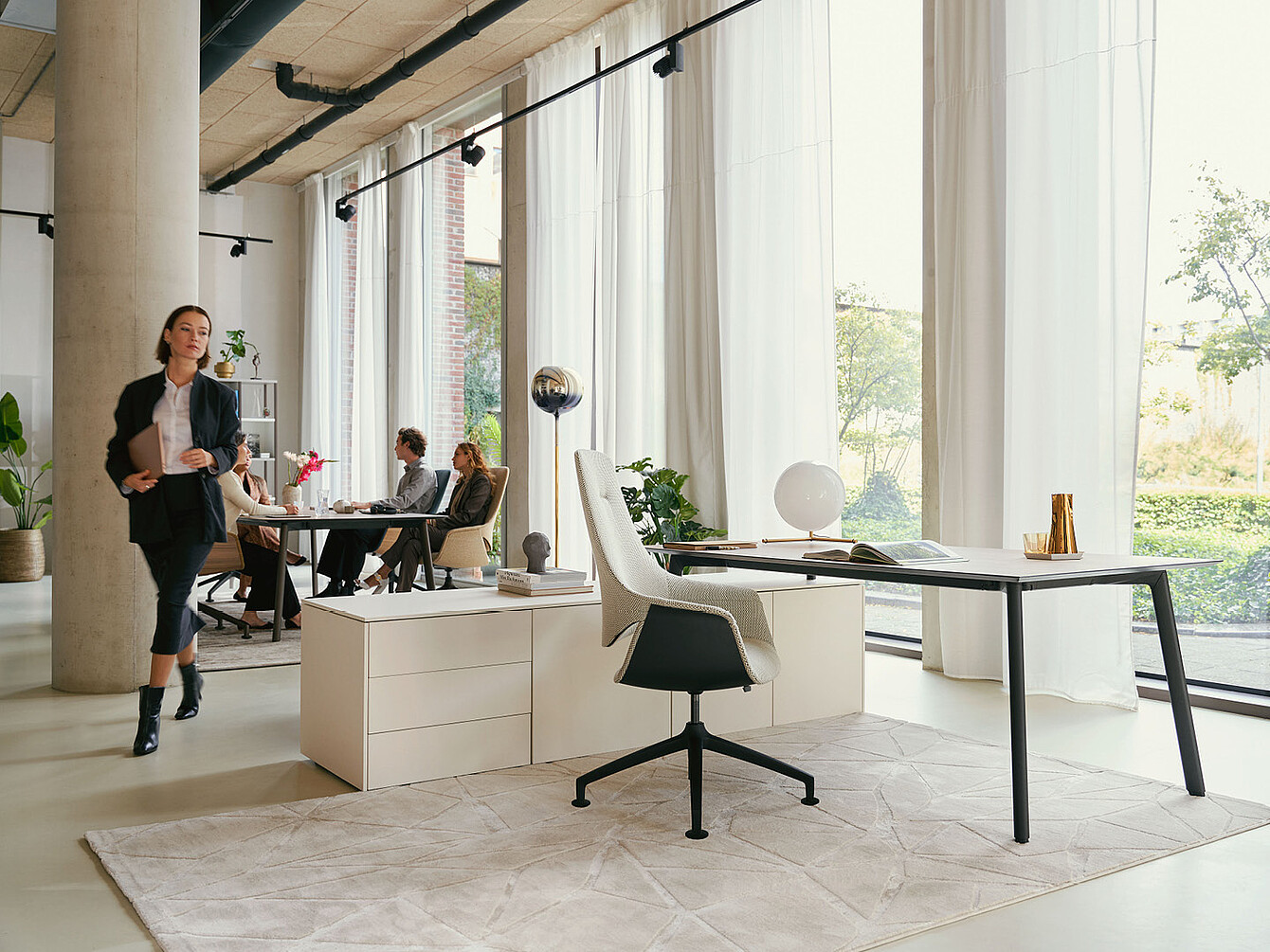 Moderne kantoorruimte met grote ramen, witte gordijnen, een bureau met een witte Sedus se:mission-draaistoel en mensen die zitten en rondlopen