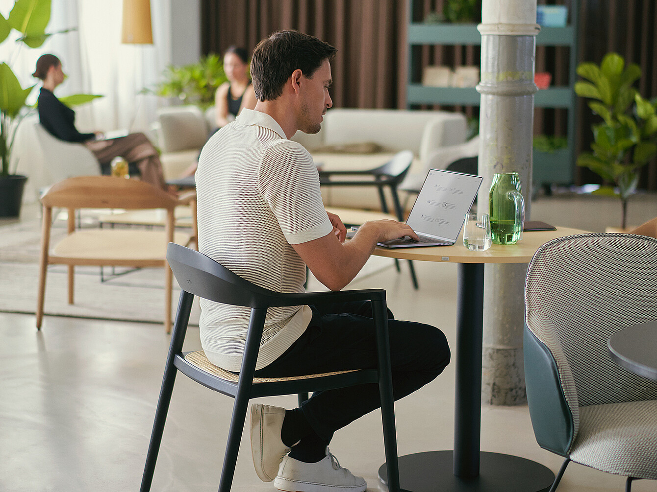Une personne assise à une table ronde dans un Work Café, utilisant un ordinateur portable avec un verre d’eau et une bouteille verte à proximité, illustrant les atmosphères variées qui stimulent l’engagement des employés.