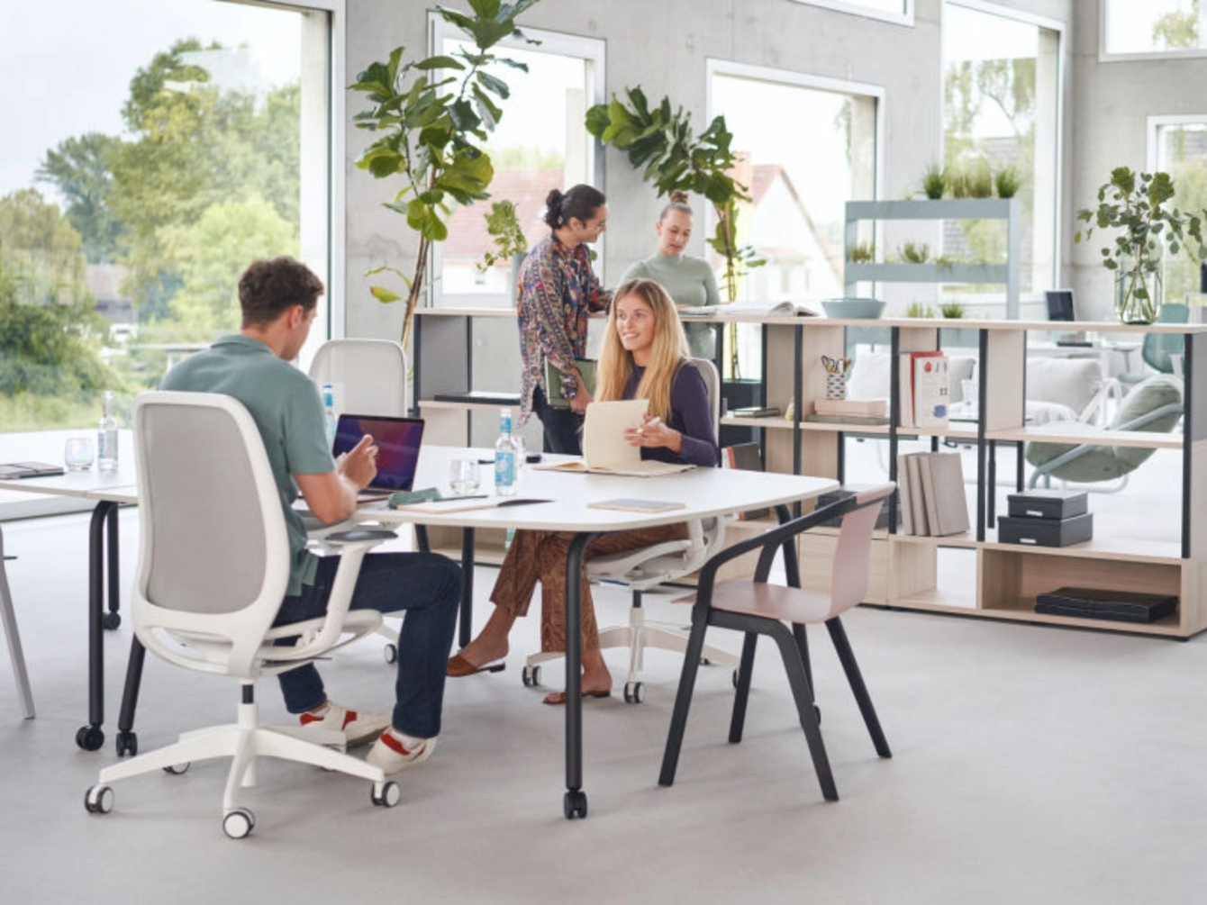 Tres personas trabajando y conversando alrededor de una mesa blanca en una oficina luminosa con grandes ventanas y plantas