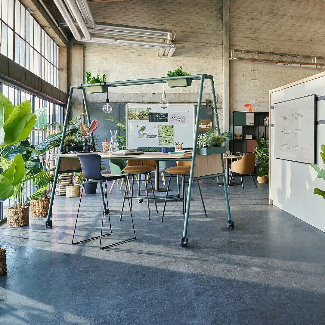 Offener, moderner Büroarbeitsplatz mit Pflanzen, Stehtischen, Sitzgelegenheiten und Whiteboards in einem loftartigen Raum mit sichtbaren Betonwänden und großen Fenstern