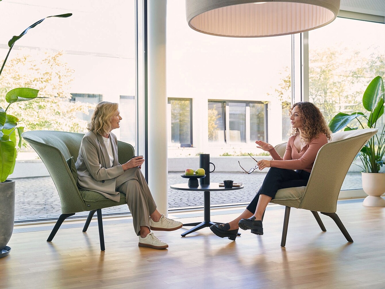 Deux femmes sont assises l’une en face de l’autre dans des fauteuils contemporains et échangent dans un espace lumineux, baigné de lumière naturelle grâce à de grandes baies vitrées et agrémenté de plantes.
