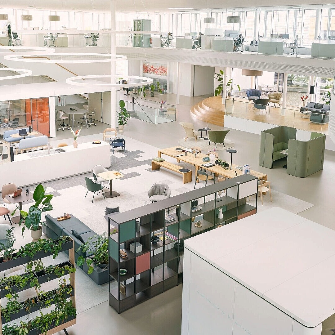 Open-plan office space with modern furniture including chairs, tables, shelving units, and plants arranged in a bright, spacious environment with large windows