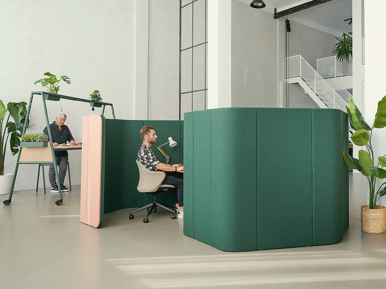 Twee werkplekken in een moderne kantooromgeving met groene akoestische scheidingswanden. Eén persoon zit aan een bureau met een lamp, terwijl een ander aan een hogere tafel met planten staat.