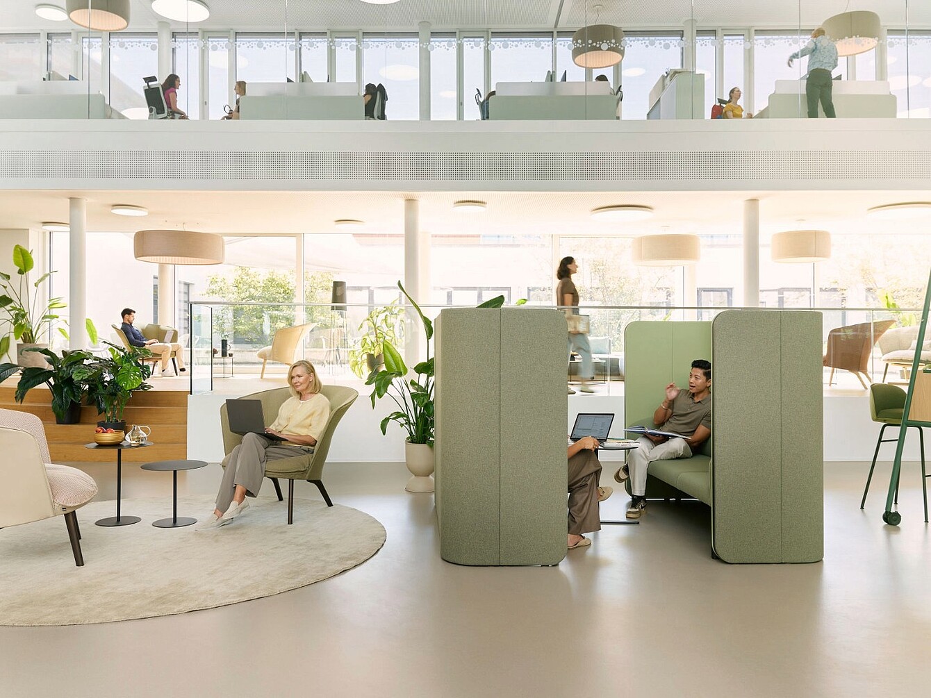 Interno di un ufficio moderno con aree a posti aperti, cabine imbottite verdi per la privacy, piante e grandi finestre che forniscono luce naturale