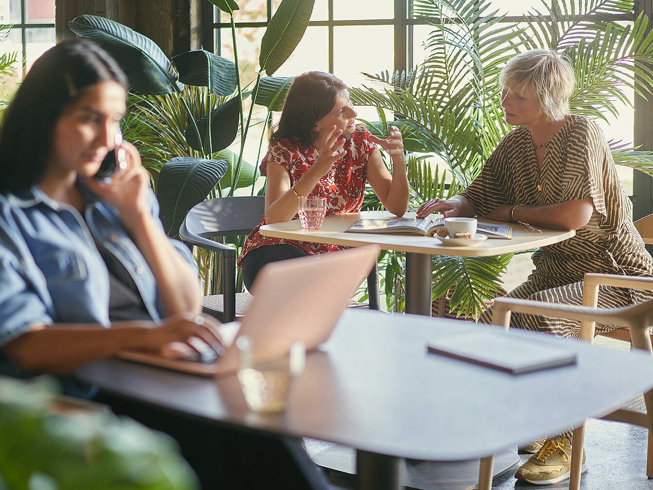 Drie vrouwen in een lichte Smart Café-kantoorruimte met grote ramen en planten; één werkt op een laptop terwijl twee anderen aan een tafel discussiëren over documenten