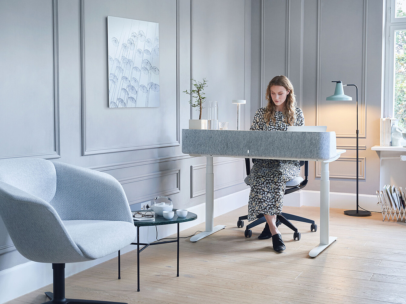 Vrouw in bloemenjurk zittend aan een modern wit Sedus-bureau in een lichtgrijze kamer met een blauwe fauteuil en staande lamp