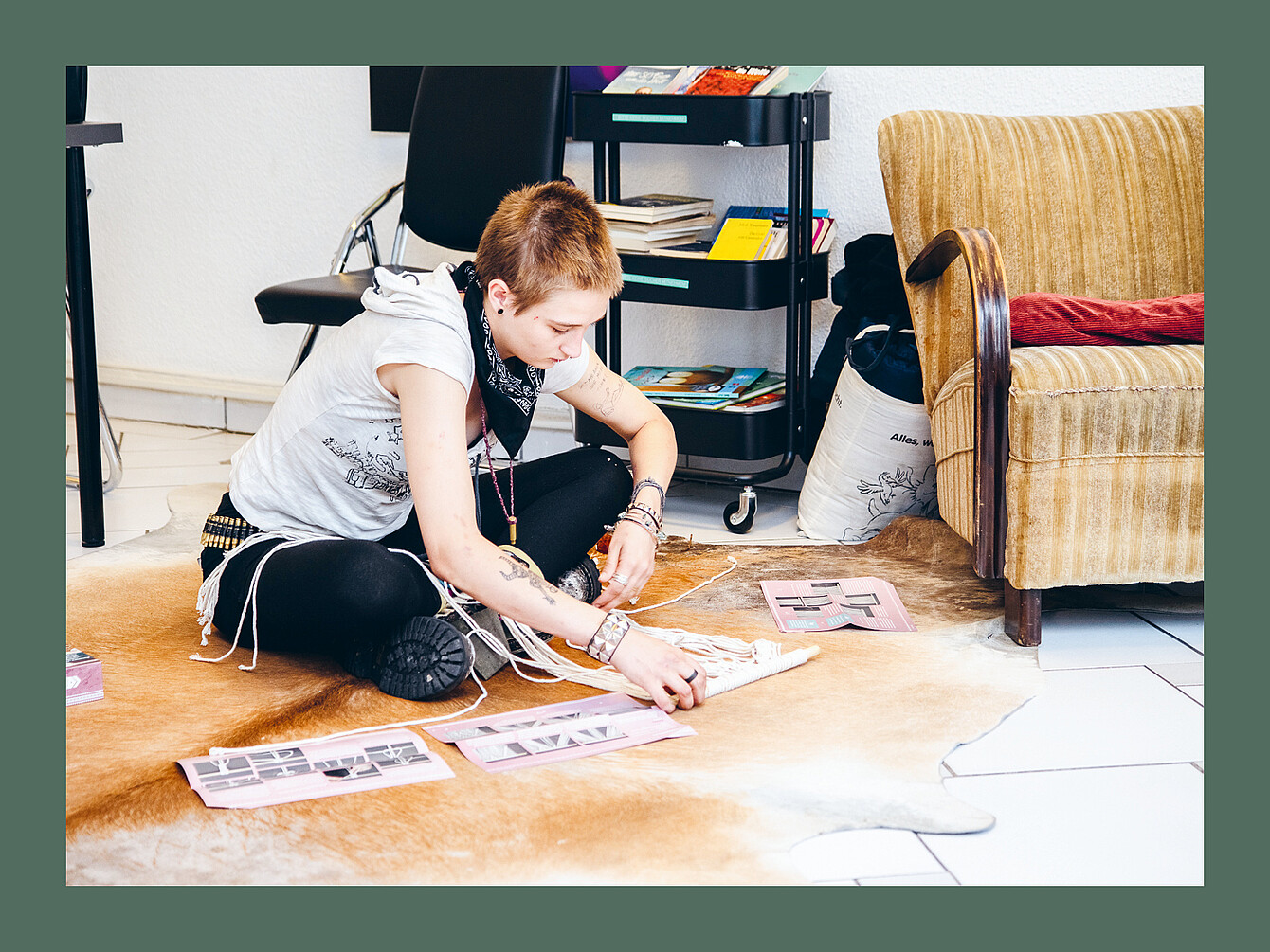 Jugendlicher sitzt auf einem hellbraunen Fellteppich, ordnet Papierbögen mit Fotos, trägt schwarzes Tuch am Hals, weiße ärmellose Kapuzenjacke und schwarze Leggings.