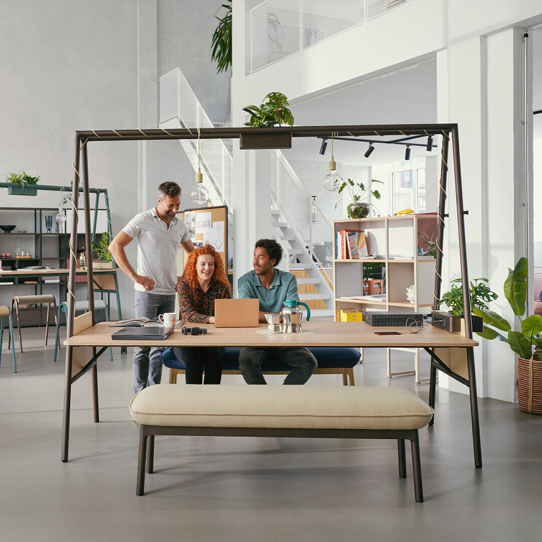 Modernes Büro mit viel Tageslicht, Pflanzen und offenen Arbeitsbereichen, drei Mitarbeiter arbeiten gemeinsam an einem Laptop, eine Person arbeitet separat am Fenster