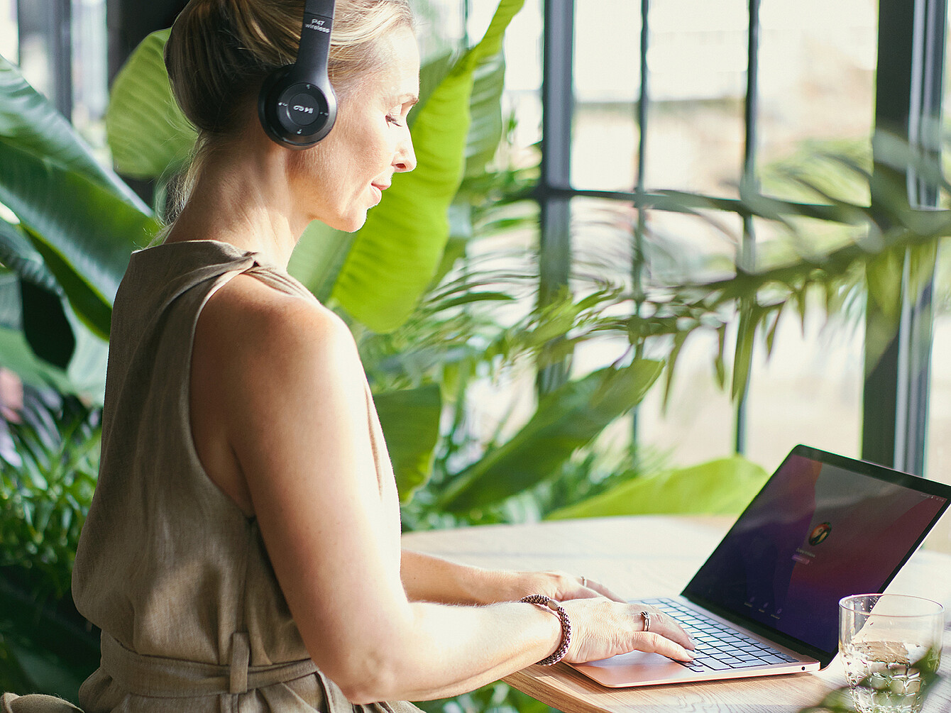 : Persona con cuffie che lavora su un laptop a un tavolo di legno circondato da grandi piante verdi in un Garden Work Café.