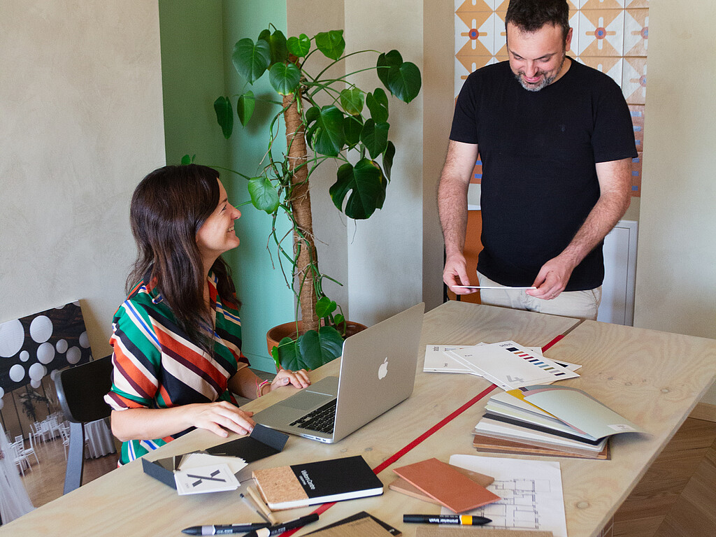 Zwei Personen arbeiten zusammen an einem Holztisch mit Laptop, Dokumenten und Notizbüchern in einem hellen Raum mit großer Pflanze