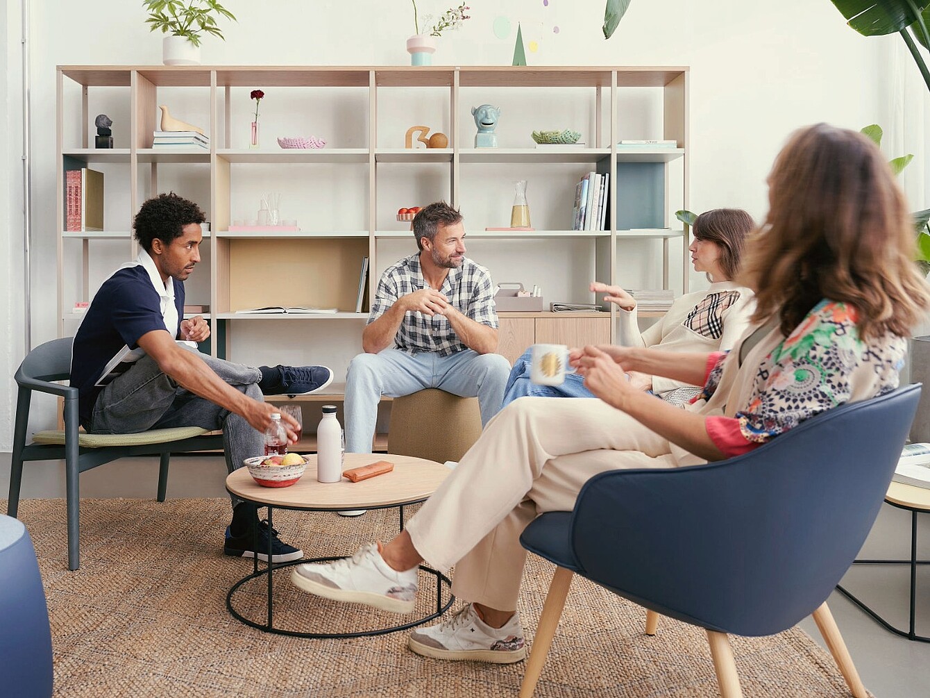 Quatre personnes assises dans un salon avec des chaises modernes et une table basse ronde devant une grande étagère avec des objets décoratifs et des livres.